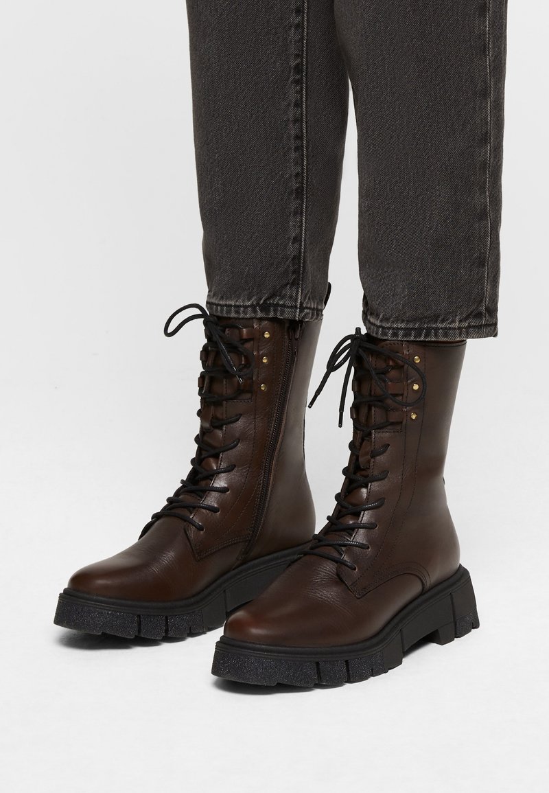 Bottes en cuir marron foncé à lacets avec semelles noires épaisses portées avec un pantalon en denim gris foncé, sur un fond neutre.