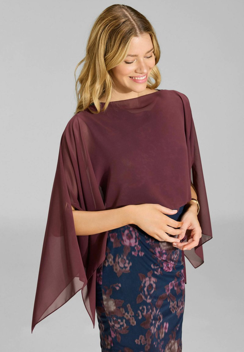 Femme aux cheveux blonds portant un haut transparent bordeaux avec des manches drapées et une jupe florale bleu marine, souriante et regardant vers le bas.