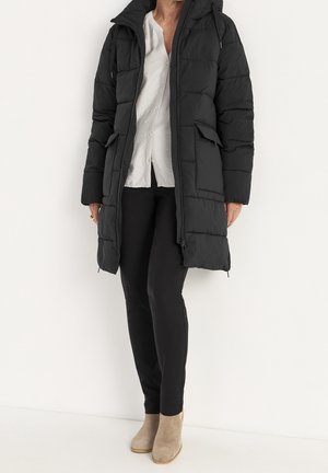 Persona che indossa un cappotto imbottito nero sopra una camicia bianca con bottoni, pantaloni neri e stivaletti beige, in piedi davanti a uno sfondo bianco.