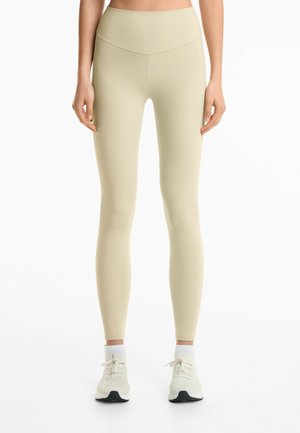 Leggings beige a vita alta con una texture liscia, forma attillata e una cintura ampia. Indossati con scarpe da ginnastica di colore chiaro.