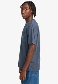 Ung man med kort, twistat hår som bär en mörkblå t-shirt och blå jeans, står sidledes och tittar på kameran.