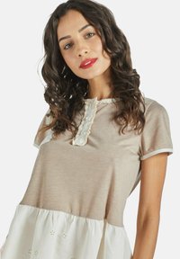 Una mujer con cabello rizado lleva una blusa beige y blanca con encaje, sonriendo ligeramente mientras mira a la cámara frente a un fondo sencillo.