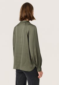Blusa verde oliva a maniche lunghe in un tessuto morbido e strutturato. Presenta un colletto classico e polsini con bottoni, orlo leggermente curvato.