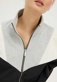 Grijs-zwarte jas met ritssluiting, voorzien van een hoge kraag en een kleurblokontwerp met een subtiele textuur en zilverkleurige ritssluiting.