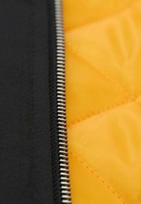 Schwarzer Stoff mit sichtbar silbernem Reißverschluss neben einer gesteppten gelben Fütterung. Enthält ein Diamantmuster und eine glatte Textur.