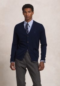 Navy cardigan made of soft knit fabric with a v-neck, three buttons, and two front pockets, paired with a striped dress shirt and tie.