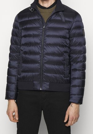 Mand iført en marineblå quiltet dunjakke over en grøn striktrøje og sorte bukser, stående foran en ensfarvet hvid baggrund.