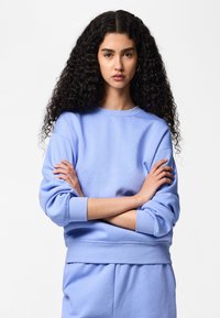 Jeune femme aux cheveux longs, bouclés et foncés, portant un sweat-shirt bleu clair et un pantalon assorti, debout les bras croisés devant un fond blanc.