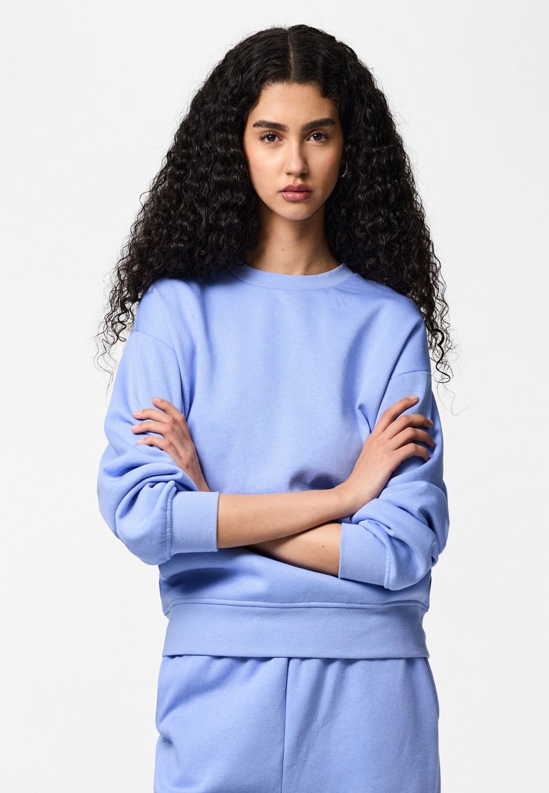 Jeune femme aux cheveux longs, bouclés et foncés, portant un sweat-shirt bleu clair et un pantalon assorti, debout les bras croisés devant un fond blanc.