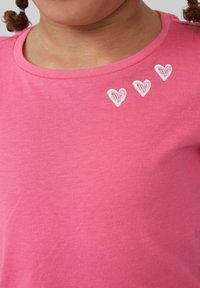 Roze katoenen T-shirt met een ronde halslijn en drie witte harten op de linkerkant van de borst. Zachte textuur, casual pasvorm.
