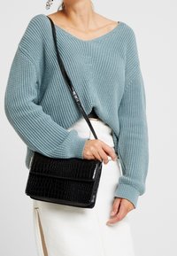 Maglione azzurro chiaro lavorato a maglia con scollo a V e texture a coste, abbinato a una borsa a tracolla nera con stampa coccodrillo.