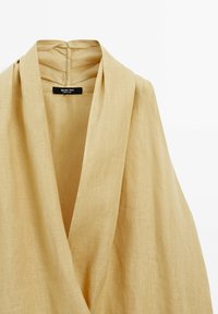 Linne omlottopp i ljus beige med djup V-ringning, mjuk textur och subtila sömmar på baksidan. Märkets etikett syns på insidan.