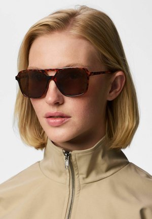 Vrouw met kort blond haar, draagt bril met schildpadprint en een beige jas met rits, tegen een effen achtergrond.