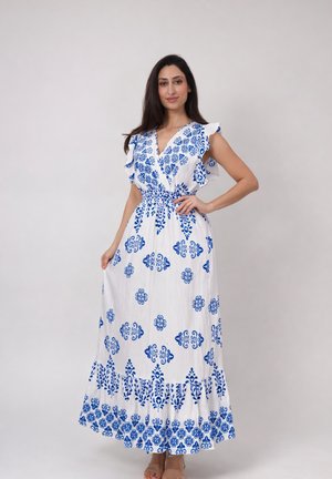 Kobieta stojąca w białej maxi sukience z niebieskimi kwiatowymi wzorami, falbaniastymi rękawami i talią podkreśloną paskiem, pozująca na jednolitym tle.