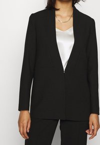 Blazer noir cintré au design élégant, avec des revers nets et une seule fermeture à l'avant. Porté sur un haut blanc en satin, avec des manches longues.