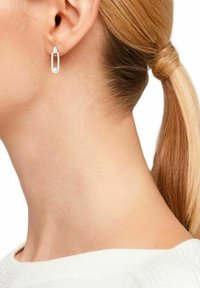 Pendientes de metal plateado en forma rectangular, con un detalle de bucle y una superficie lisa, llevados en la oreja de un modelo con cabello claro.