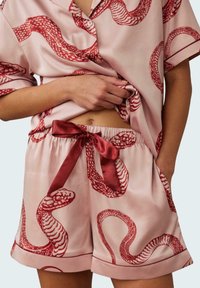 Satin loungewear set featuring a loose-fit shirt and shorts, pale pink with red snake patterns, and a satin tie on shorts.