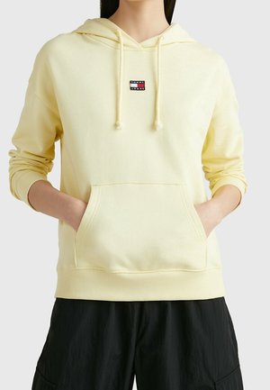 Personne portant un sweat à capuche jaune pâle avec poche avant et logo "Tommy Jeans", associé à un pantalon noir ample, mains dans les poches.