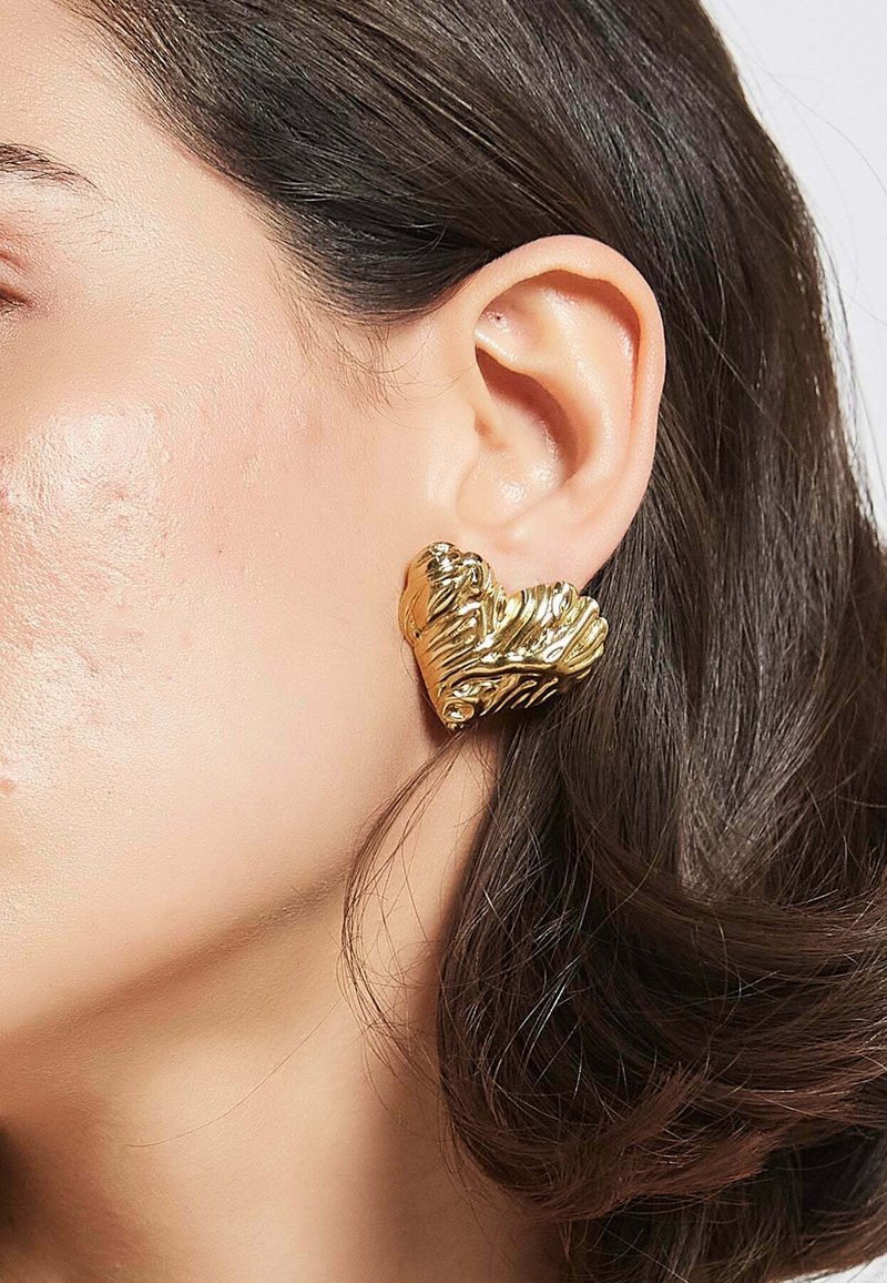 Guldörhänge i hjärtform med en strukturerad yta, som har svängande detaljer och en glansig finish, bärs på en mannekängöras.