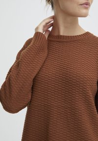 Suéter marrón de punto con un patrón texturizado, cuello acanalado y mangas largas. Primer plano del cuello y área del hombro.