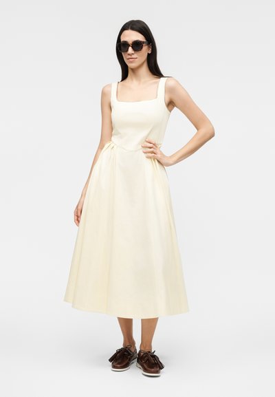 Γυναίκα με μπεζ αμάνικο μίντι φόρεμα, σκοτεινά γυαλιά ηλίου και καφέ μοκασίνια με φουντίτσες, στέκεται με το ένα χέρι στη μέση.