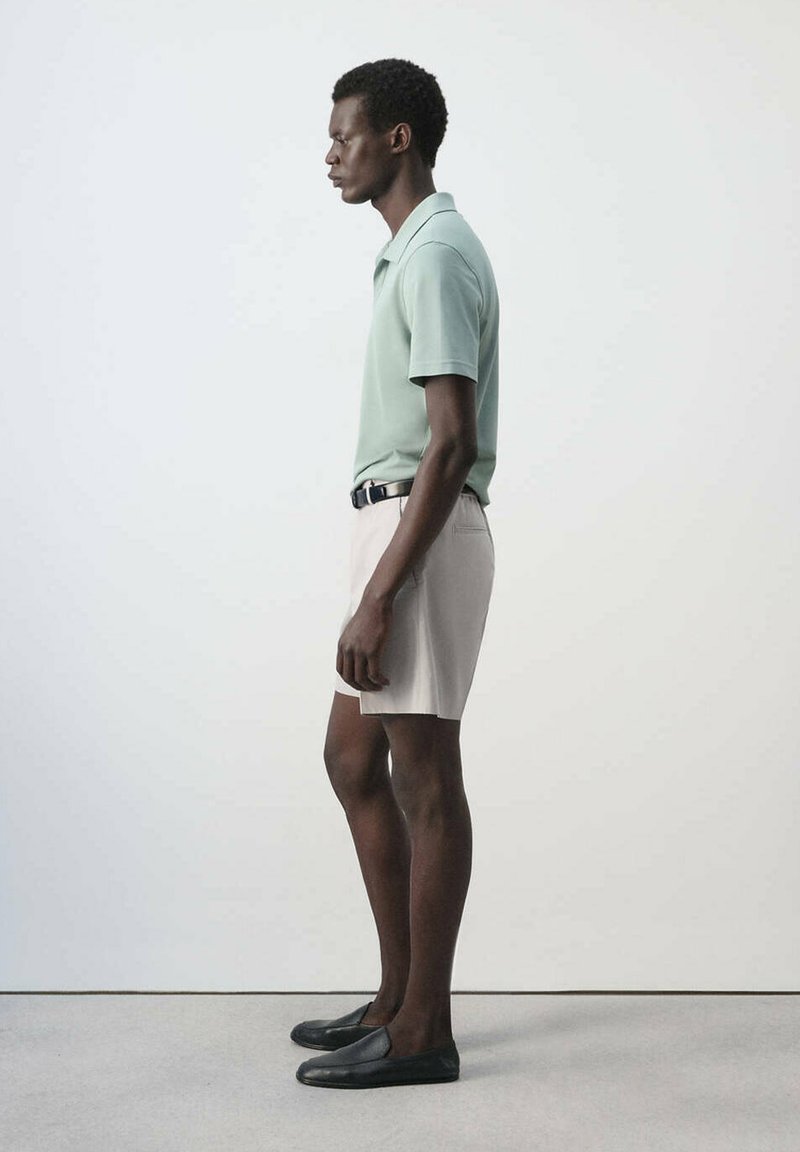 Man standing in profile wearing a light green polo shirt, beige shorts with a black belt, and black slip-on shoes against a plain background.