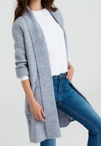Cardigan azzurro chiaro con un motivo a maglia testurizzato, apertura frontale e maniche lunghe. Presenta tasche laterali ed è abbinato a una maglietta bianca e jeans blu.