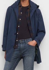 Námornícká nepromokavá bunda s kapucí, která má zapínání na zip a knoflíky. Nositelná přes kostkovanou košili a tmavé tričko, doplněná modrými džíny.