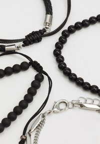 Joyería negra que incluye cuero trenzado, cuentas redondas y un broche de plata. Varias texturas y formas crean una impactante exhibición monocromática.