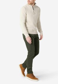 Cremfarbener Strickpullover mit einem halben Reißverschluss-Kragen, kombiniert mit dunkelgrünen Cordhosen und braunen Wildleder-Schnürschuhen.