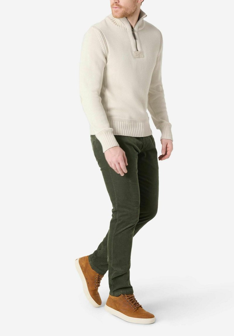 Cremfarbener Strickpullover mit einem halben Reißverschluss-Kragen, kombiniert mit dunkelgrünen Cordhosen und braunen Wildleder-Schnürschuhen.