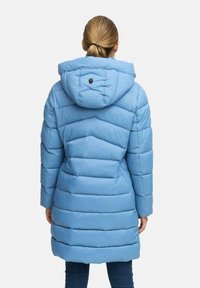 Vrouw die wegkijkt in een lichtblauwe, gewatteerde lange pufferjack met capuchon, gecombineerd met donkerblauwe jeans.