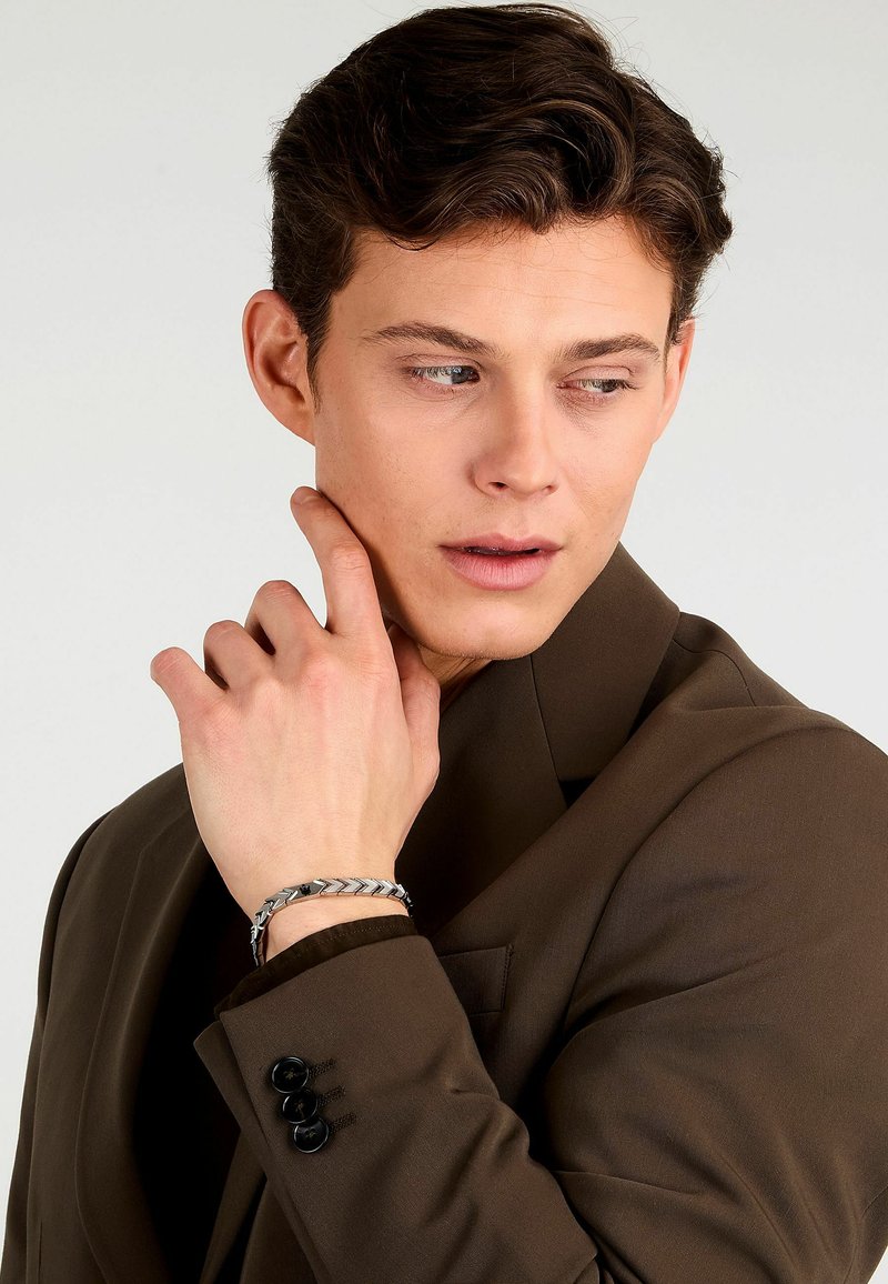 Bracciale in argento con design a zig-zag indossato su una mano, abbinato a una giacca marrone con bottoni neri.