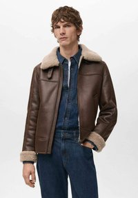 Giacca di pelle marrone con colletto e polsini in shearling. Presenta una chiusura con zip, una texture liscia e una forma sagomata. Indossata sopra una camicia di denim.