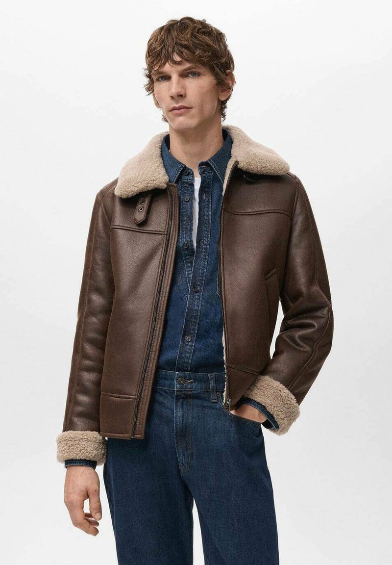 Giacca di pelle marrone con colletto e polsini in shearling. Presenta una chiusura con zip, una texture liscia e una forma sagomata. Indossata sopra una camicia di denim.