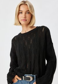 Schwarzer Strickpullover mit einem strukturierten Muster aus vertikalen Linien und offenen Maschen. Lockere Passform mit weiten Ärmeln und rundem Ausschnitt.