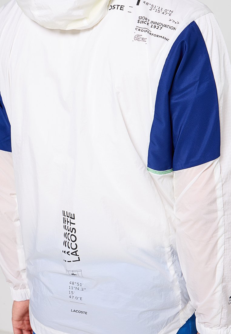 Veste légère blanche avec des manches bleues, dotée d'une capuche, d'une marque imprimée et de coordonnées géographiques dans le dos. Tissu texturé.