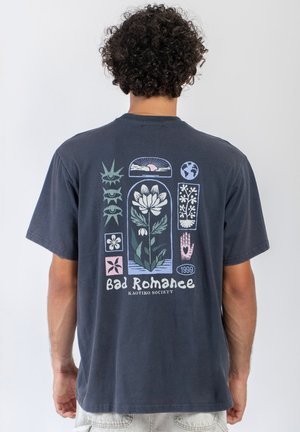 Mężczyzna w ciemnej koszulce z kwiatowymi i symbolicznymi wzorami, w tym oczy, słońce, ziemia, ręka oraz tekstem "Bad Romance Kaotiko Society 1999" na plecach.