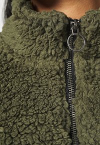 Chaqueta de forro polar verde oliva con superficie texturizada, cuello alto y cierre frontal con una lengüeta circular.