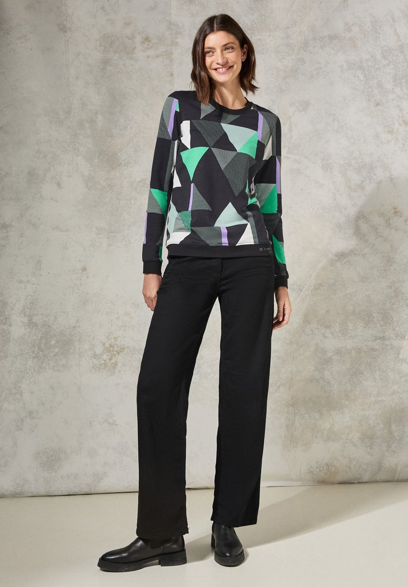 Maglia a maniche lunghe con una base nera decorata con motivi geometrici verde, turchese e viola. Abbinata a pantaloni larghi neri e stivaletti alla caviglia.