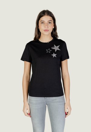 Mladá žena v černém tričku s třemi stříbrnými hvězdami na levé straně hrudi a světle šedých džínách stojící na jednobarevném pozadí.