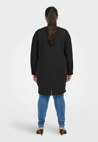 Tunica nera a maniche lunghe con orlo spaccato, abbinata a jeans aderenti blu e scarpe col tacco nere, vista da dietro.