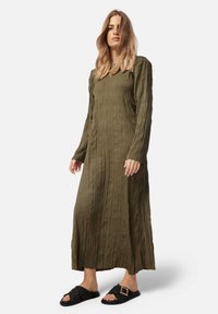 Lungo vestito verde oliva con un design texturizzato e plissettato, dotato di maniche lunghe e una vestibilità comoda, abbinato a sandali neri texturizzati senza chiusura.