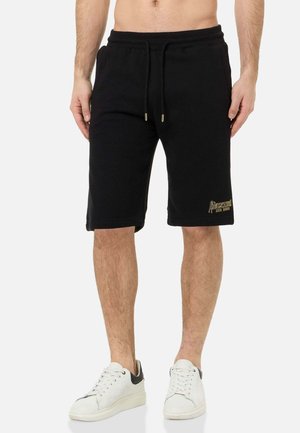 Mann trägt schwarze Shorts mit Kordelzug und goldenem Logo am linken Bein und weiße Sneakers mit schwarzen Fersenakzenten.
