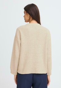 Šviesiai rudas megztinis su ilgomis rankovėmis, raukšlėta tekstūra ir laisva kirpimo linija. Nugaros dalyje yra paprastas apvalus iškirpimas ir jokių matomų užsegimų.