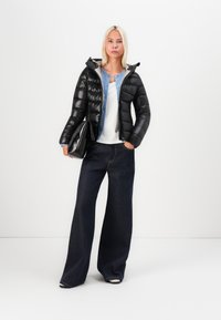 Svart pufferjakke med horisontalt quiltet tekstur, grå hettefôr, kombinert med en lyseblå denimskjorte, hvit topp og vide, mørke jeans.