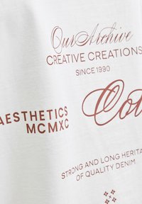 Hvid bomulds-T-shirt med rød trykt tekst, inklusive "Our Archive," "Aesthetics MCMXC," og "Strong and Long Heritage of Quality Denim."