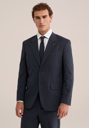 Man die een donkerblauw pak draagt met een wit overhemd en een donkere stropdas, staand tegen een lichtbeige achtergrond.