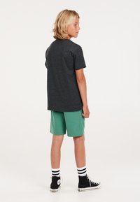 Zwarte t-shirt met korte mouwen, groene shorts, zwarte hoge sneakers met witte accenten, wit en zwarte gestreepte sokken, staand van achteren.
