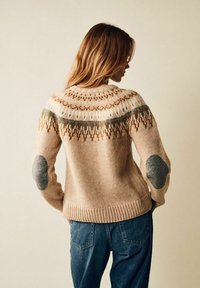 Stickad tröja i beige med ett mönstrat ok i brunt och turkos. Har grå armbågsremmar och en ribbad nederkant, kombinerad med blå jeans.